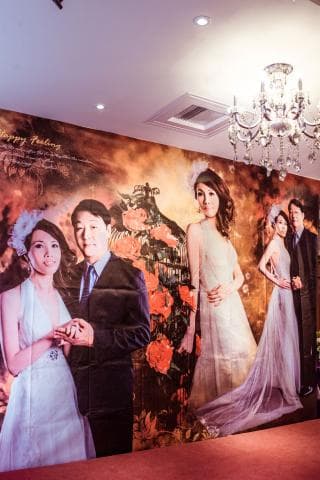 這張婚禮的照片是一個婚禮場地的佈置，背景是一幅大大的婚紗照，上面有兩對新人，一個穿著白色婚紗，一個穿著西裝，他們手牽手站在一起，看起來很幸福。