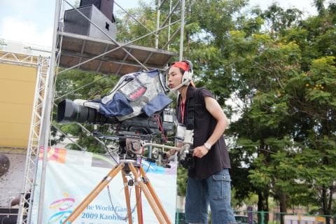 這張照片是一個男子操作著大型的攝影機，他戴著耳機，表情專注，似乎正在拍攝比賽，畫面充滿動感。