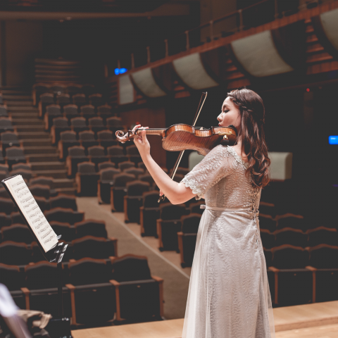 這張照片是一個年輕女子在舞台上演奏小提琴。她穿著優雅的白色連衣裙，頭髮披肩，表情專注，看起來很有魅力。她演奏的小提琴是照片中最顯眼的部分，它的曲線優美，泛著溫暖的色澤，吸引著觀者的目光。