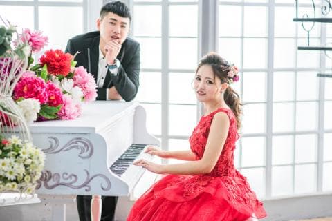 這張「婚紗照」的照片是一對新人站在白色鋼琴前，新娘穿著紅色婚紗，新郎穿著黑色西裝，背景是白色窗戶。