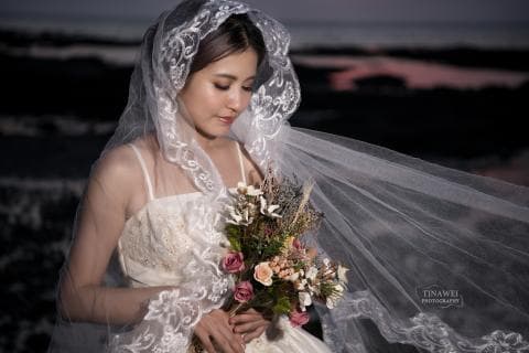 這張婚紗照是一個新娘穿著蕾絲婚紗，頭戴頭紗，手捧花束，站在海邊的畫面。