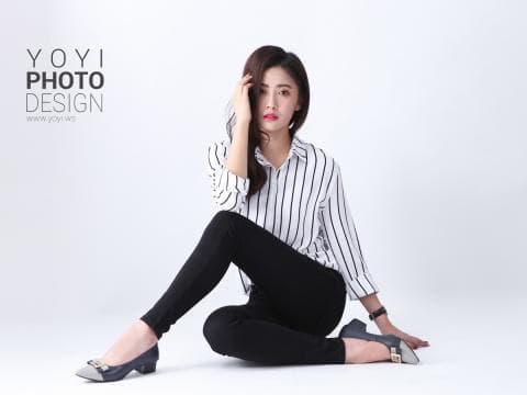 這張「時尚攝影」的照片是一個穿著白襯衫和黑色褲子的年輕女人，她坐在白色背景上，一隻手放在頭上，表情自信。