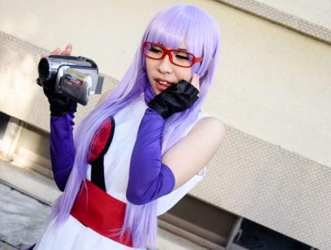 這張照片是一名紫色頭髮的女孩，她戴著紅色眼鏡，手拿著一臺攝像機，她正看著鏡頭微笑。