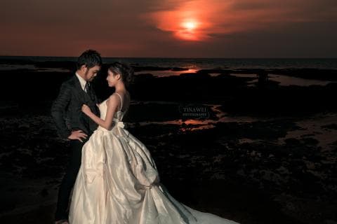 這張照片是一對新人站在海邊，夕陽西下，背景是紅色的天空和黑色的岩石。新郎穿著黑色的西裝，新娘穿著白色的婚紗，他們深情地互相凝視，顯得非常恩愛。