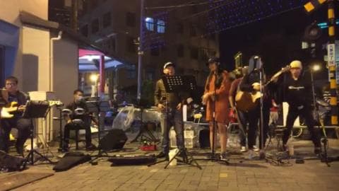 這張「街頭表演」的照片是一群人在街道上表演，他們有吉他、鼓、麥克風，以及一個女歌手。他們似乎在晚上表演，周圍有一點燈光。