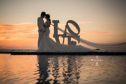 這張照片是一對新人在夕陽下，面對著巨大的 LOVE 字樣，新娘披著長長的頭紗，新郎輕輕摟著新娘的腰，看起來幸福又甜蜜。