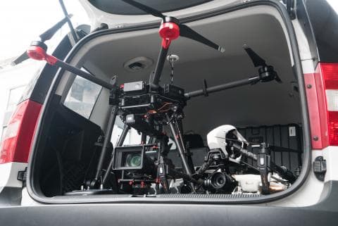 這張照片是一輛汽車的後車廂，裡面放著一臺大型無人機和一部相機。無人機的機身很大，有四個螺旋槳，機身下方安裝著一臺攝影機。相機是專業的，有一個大的鏡頭。無人機和相機一起構成了一個強大的影像採集系統，可以用於拍攝航拍影片或照片。