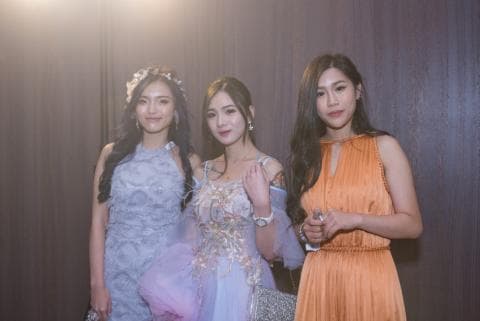 這張「三個穿著禮服的年輕女性」的照片是，她們都笑著，站在木牆的背景前。她們的髮型和服裝都很正式。