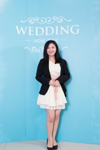 這張照片是一個穿著白色洋裝和黑色西裝外套的年輕女人，她站在藍色背景的前面，背景上寫著「WEDDING」。