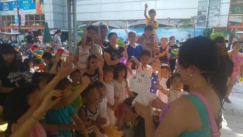 這張照片是很多大人和小孩聚集在一起的照片，最重要的是照片裡面的女性拿著一個白色大貓頭的玩偶，她正準備與小朋友玩遊戲。