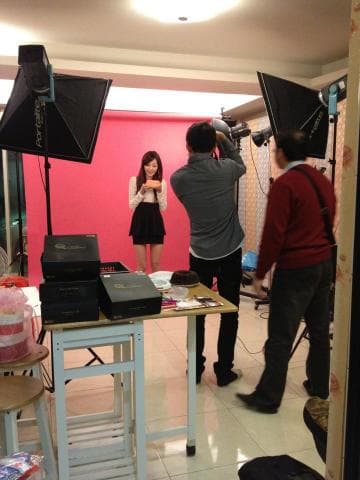 這張「攝影棚拍攝」的照片是，一個攝影師正在拍攝一位穿著白色上衣和黑色裙子的女生，旁邊還有攝影棚的工作人員，背景是一片粉色的牆。