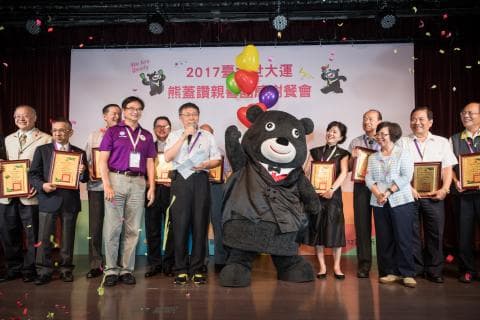 這張照片是一群人站在大型黑熊玩偶前面，黑熊玩偶穿著西裝，戴著領帶，看起來很紳士。