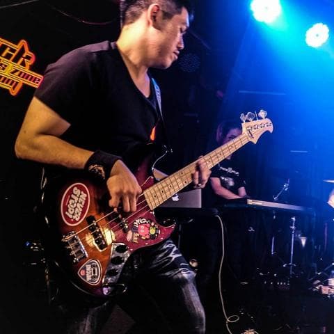 這張照片是一個男子彈奏低音吉他的照片。他穿著黑色T恤，戴著帽子，專注地彈奏著低音吉他。他身後有一個人，但他並不在照片的中心位置。低音吉他上有許多裝飾貼紙，看起來很酷。