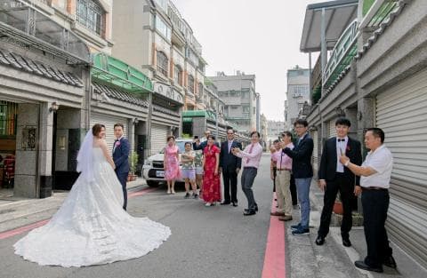 這張照片是一對新人正在街道上拍婚紗照，新郎穿著西裝，新娘穿著白色婚紗，他們旁邊站著許多親朋好友，大家都在笑著祝福他們。