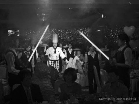 這張照片是一群人在一個宴會廳裡吃飯，其中一位廚師身穿白圍裙，正向大家展示他的拿手好菜。