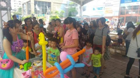 這張照片是一個街頭表演，表演者正在用氣球製作各種動物造型，吸引許多大人和小孩圍觀。