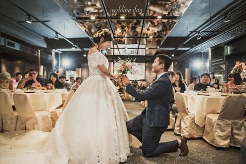 這張婚禮的照片是新郎跪在地上向新娘求婚，新娘穿著白色婚紗，新郎則穿著深色西裝，背景是婚宴的場地，佈置著許多桌椅和花朵。