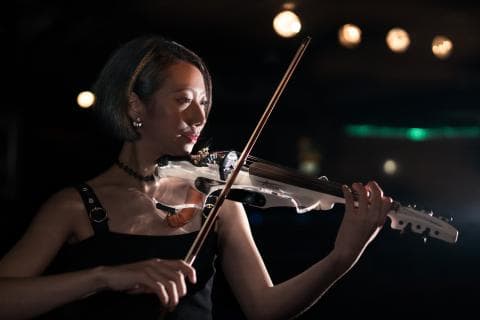 這張照片是一個年輕女子正在演奏電小提琴。她專注的眼神和熟練的手指動作，展現了對音樂的熱愛。她手上的電小提琴，散發出銀色的光澤，在黑暗中顯得格外耀眼。