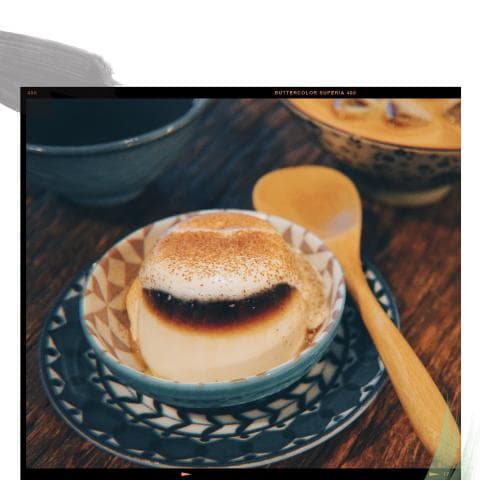 這張「布丁甜點」的照片是一碗上面撒了肉桂粉的布丁，旁邊放著一把木勺和一個裝滿茶的碗。