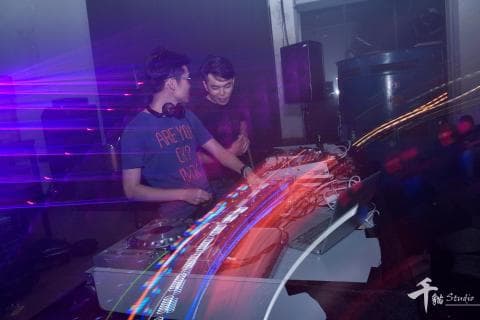 這張照片是兩個年輕人在一個音樂工作室裡，其中一個正在操作混音臺，而另一個則在一旁觀看。他們周圍充滿著五顏六色的燈光，營造出熱鬧的氛圍。