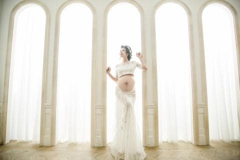 這張照片是一位身穿白色蕾絲洋裝的孕婦，她站在三扇窗戶前，露出幸福的笑容，看起來非常美麗動人。