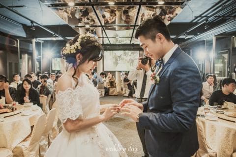 這張婚禮的照片是一個新郎新娘在婚宴上交換戒指的畫面，新娘穿著白色婚紗，新郎穿著深藍色西裝，他們都很開心。