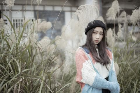 這張「女孩站在草叢中」的照片是一個穿著毛衣的女孩，她站在一大片高高的草叢中，背景是一些建築。