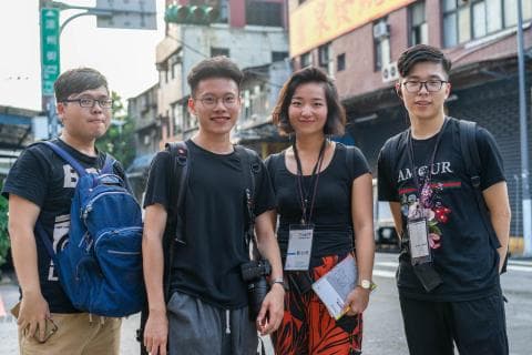 這張「四個朋友站在一起」的照片是，他們都穿著休閒服，其中一人穿著一件印有花紋的黑色 T 恤，背景是一條街道。