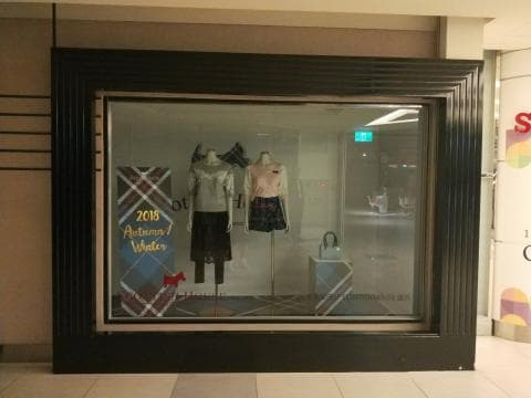 這張照片是一家商店的櫥窗，裡面展示了兩件女裝，一件是白色上衣搭配黑色裙子，另一件是粉色上衣搭配格紋短裙。櫥窗裡還有一個手提包和一個紅色的小狗模型，最顯眼的則是櫥窗中央的「2018 Autumn/Winter」標語。