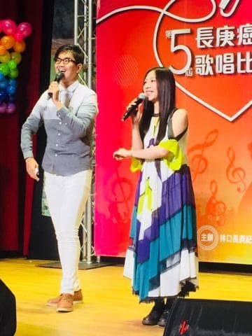 這張照片是一個舞臺，兩個表演者在唱歌。左邊的男生穿著淺色襯衫和白色褲子，拿著麥克風。右邊的女生穿著鮮豔的裙子，也拿著麥克風。