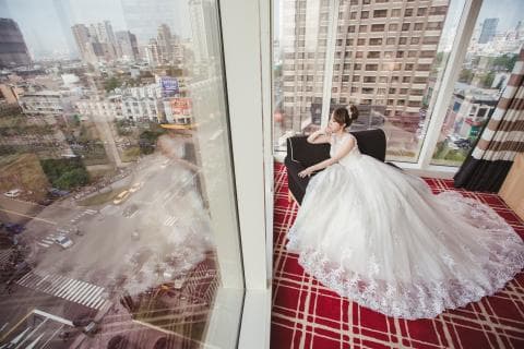 這張照片是一個穿著白色婚紗的新娘坐在窗邊的沙發上。她優雅地看著窗外的城市風景。