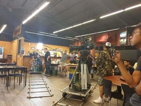 這張「電影拍攝現場」的照片是，一群人在一個空曠的房間裡拍攝電影，攝影師正在使用專業攝影機拍攝，演員們在鏡頭前表演，場景佈置簡陋，但氛圍熱鬧。