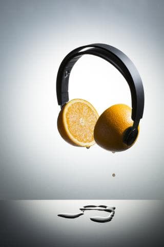 這張「耳機和橘子」的照片是一副黑色耳機，上面放著兩個橘子切片，橘子正在滴汁，掉落在光滑的表面上。