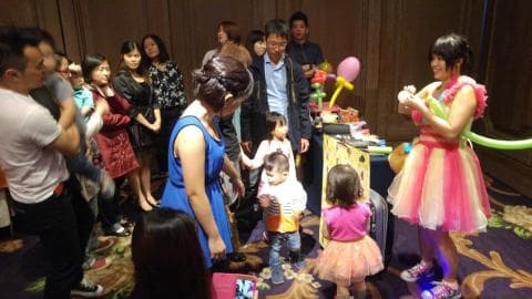 這張照片是許多人在一個房間裡慶祝，其中一個穿著粉紅色禮服的女性正在為小朋友們表演氣球藝術。