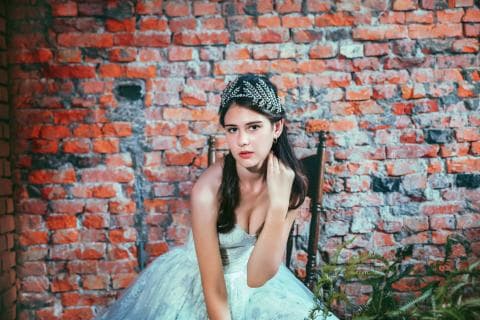 這張「婚紗照」的照片是一個穿著白色婚紗的女孩，她戴著皇冠坐在紅磚牆前，她的表情看起來很幸福。