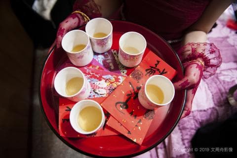 這張照片是擺滿了茶杯的紅色託盤，上面的茶杯裝滿了茶，一旁放著紅色的喜帖，看起來像是在婚禮上喝茶的儀式。