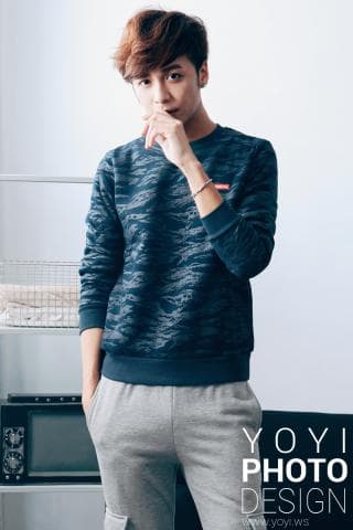 這張照片是一個年輕男子穿著藍色迷彩上衣和灰色運動褲，他用手捂著嘴巴，顯得有些害羞。