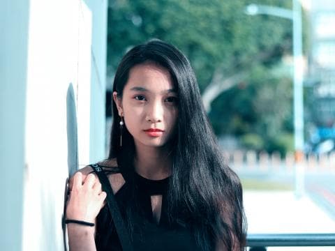 這張「年輕女子」的照片是一個穿著黑色背心，長髮披肩的女孩，她站在白色牆壁旁，眼神望向鏡頭，顯得冷靜沉穩。