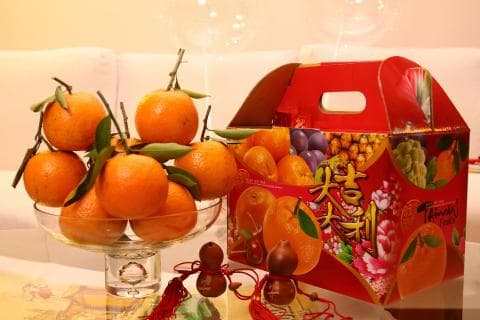 這張「水果和禮盒」的照片是一些橙子和一個紅色禮盒。橙子放在一個玻璃盤子裡，禮盒放在桌上。禮盒上有中國字和一些水果的圖案。