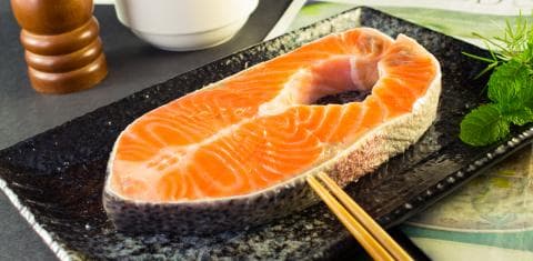 這張「鮭魚」的照片是一片新鮮的鮭魚排，放在黑色盤子上，旁邊還有一雙筷子和一些綠色的葉子。