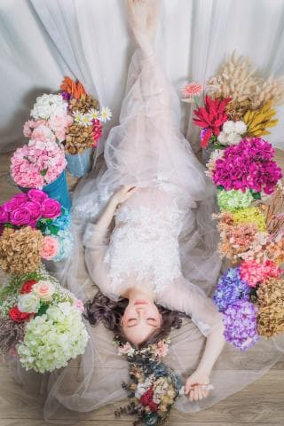 這張照片是一個穿著婚紗的女子，她躺在地上，頭上戴著花環，四周圍繞著各種花卉，呈現出浪漫且幸福的氛圍。