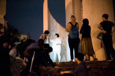 這張照片是一個在晚上戶外舉行的派對，一群朋友圍繞在一個現代建築物旁邊，其中一個穿著白色衣服的女孩站在中間，顯得格外突出。