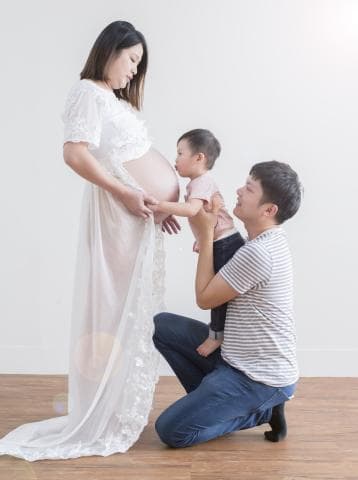 這張照片是一對夫妻，丈夫跪在懷孕的妻子面前，並且看著妻子隆起的肚子，他們的小孩則伸手觸摸著媽媽的肚子。