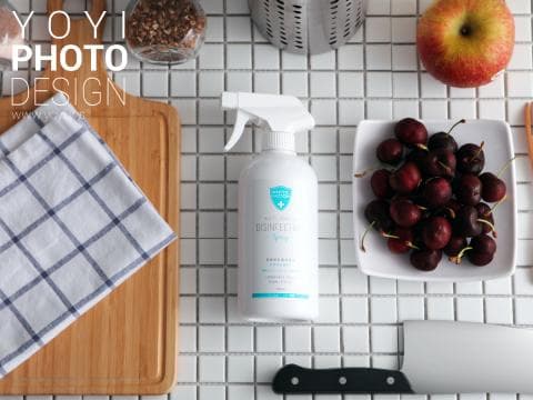 這張「廚房用品」的照片是廚房裡一個木質砧板、一瓶消毒噴霧、一塊格紋布巾、一個裝滿紅櫻桃的白色碟子、一個蘋果和一把刀。