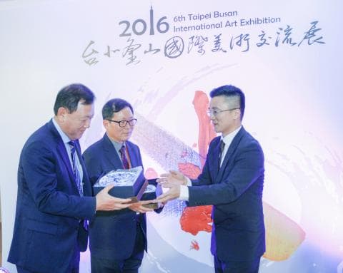 這張「2016年臺北釜山國際藝術展」的照片是三位男士站在展覽的背景前，其中兩位男士手持獎牌，照片的背景是展覽的標誌和裝飾。