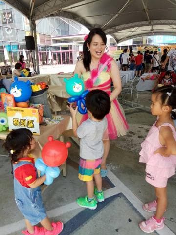 這張「親子互動」的照片是一個女人在教小朋友做氣球動物。她穿著粉紅色洋裝，手上拿著藍色的氣球動物。旁邊的小朋友穿著彩色衣服，看起來很開心。