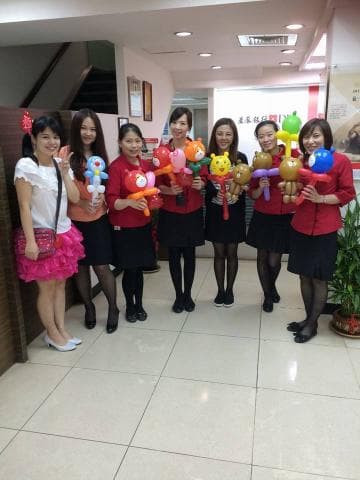 這張照片是六位女性站在一起，她們都穿著紅色上衣和黑色裙子，手中拿著氣球。她們看起來很開心，似乎在參加一個活動。