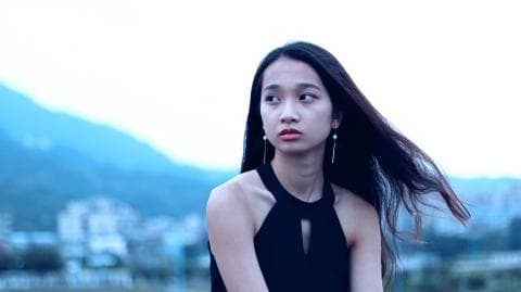 這張「年輕女子」的照片是，她穿著黑色吊帶背心，頭髮飄逸，背景是模糊的城市風景。