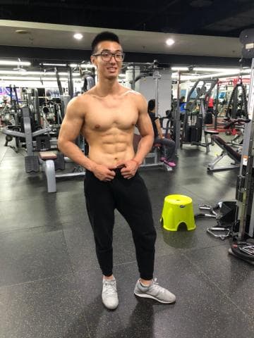 這張「健身房」的照片是一個年輕男子，他穿著黑色運動褲，站在健身房裡，他的身形健壯，可以看出他經常鍛鍊。他戴著眼鏡，面帶笑容，看起來很自信。