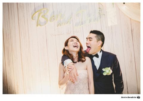 這張婚禮的照片是一對新人在木質背景前開心大笑，新郎穿著黑色西裝，新娘穿著米色禮服。